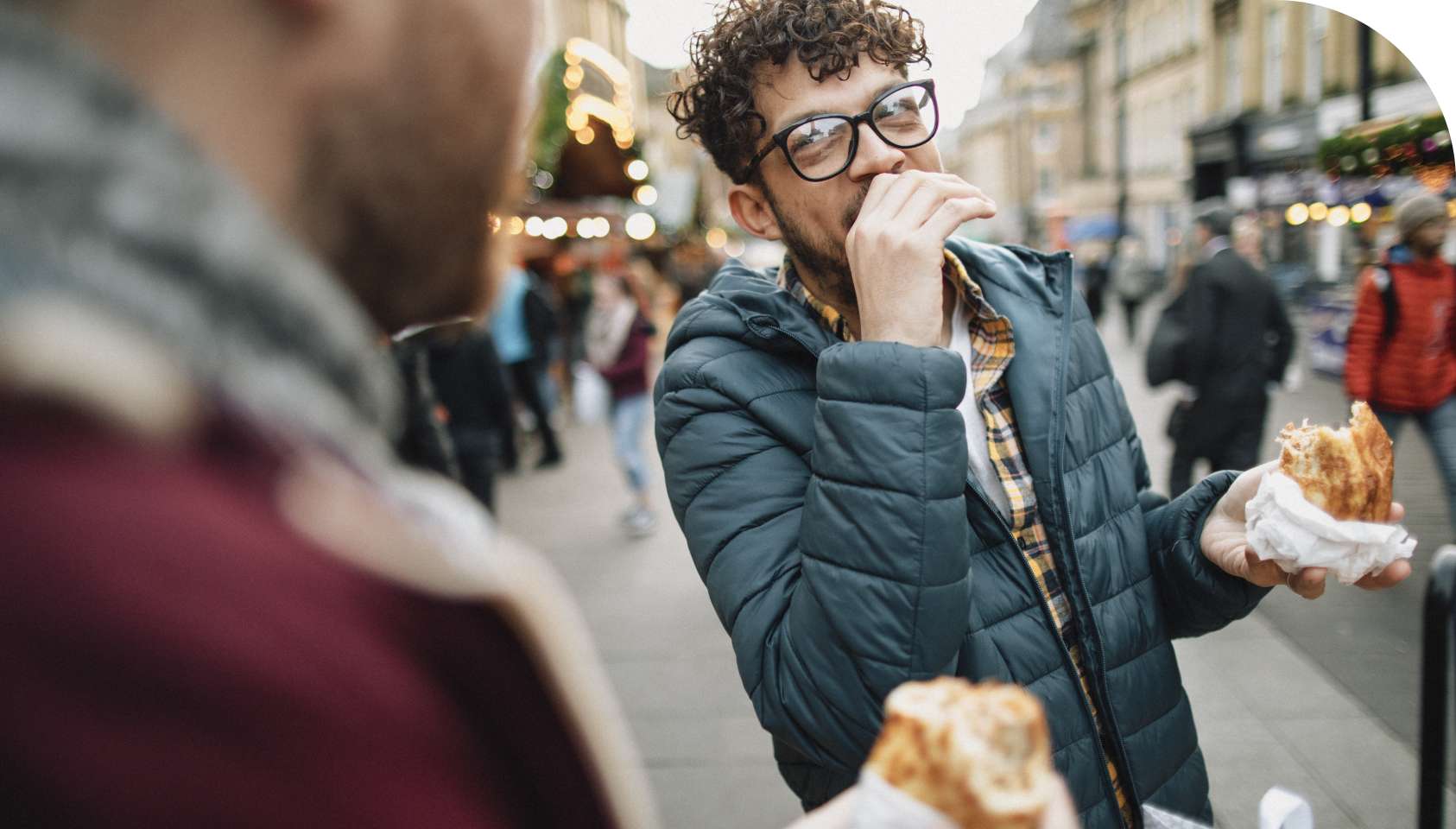 A young man wearing black framed glasses and a blue coat is at an outdoor event in the city eating a sandwich while talking to a friend